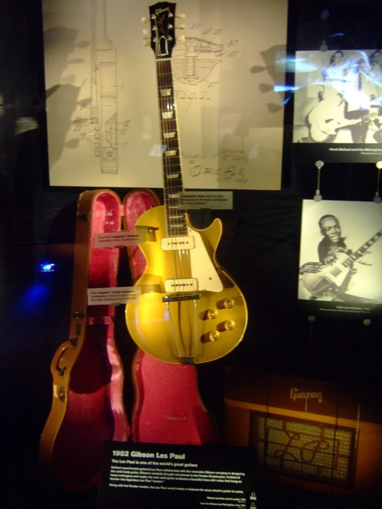 Золотистая гитара Gibson Les Paul 1952 года в музейной витрине, рядом открытый футляр с розовой подкладкой, за ней патентный чертёж