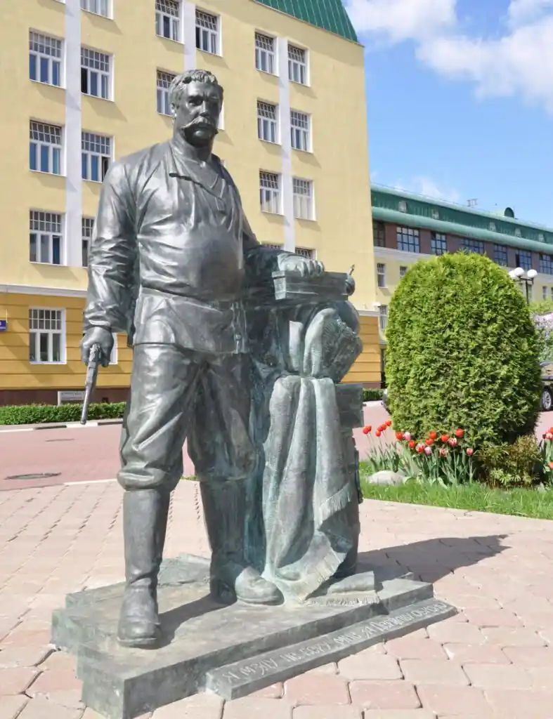 Бронзовый памятник: усатый мужчина в рубахе и сапогах с пистолетом в руке стоит у ящиков и свёрнутого ковра, на постаменте надпись