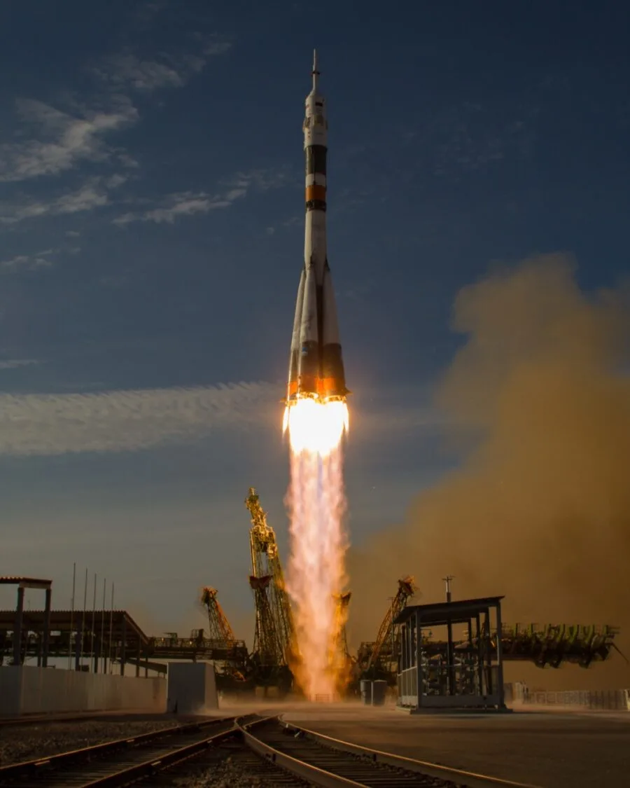 Ракета «Союз» поднимается со стартового стола на столбе огня, по сторонам решётчатые мачты, справа облако бурого дыма, вечернее небо