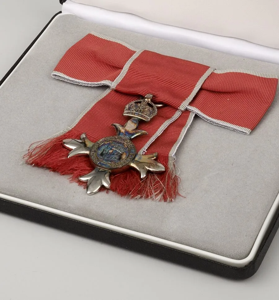 Серебристый знак ордена Британской империи степени MBE на красной ленте с серой каймой, лежащий в чёрном футляре с серой бархатной вставкой