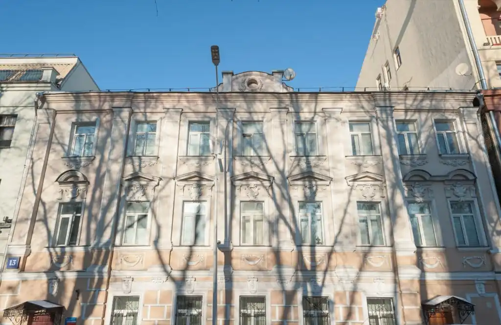 Трёхэтажный дом с лепниной на фасаде, на углу табличка с номером 13, по стене лежат тени ветвей